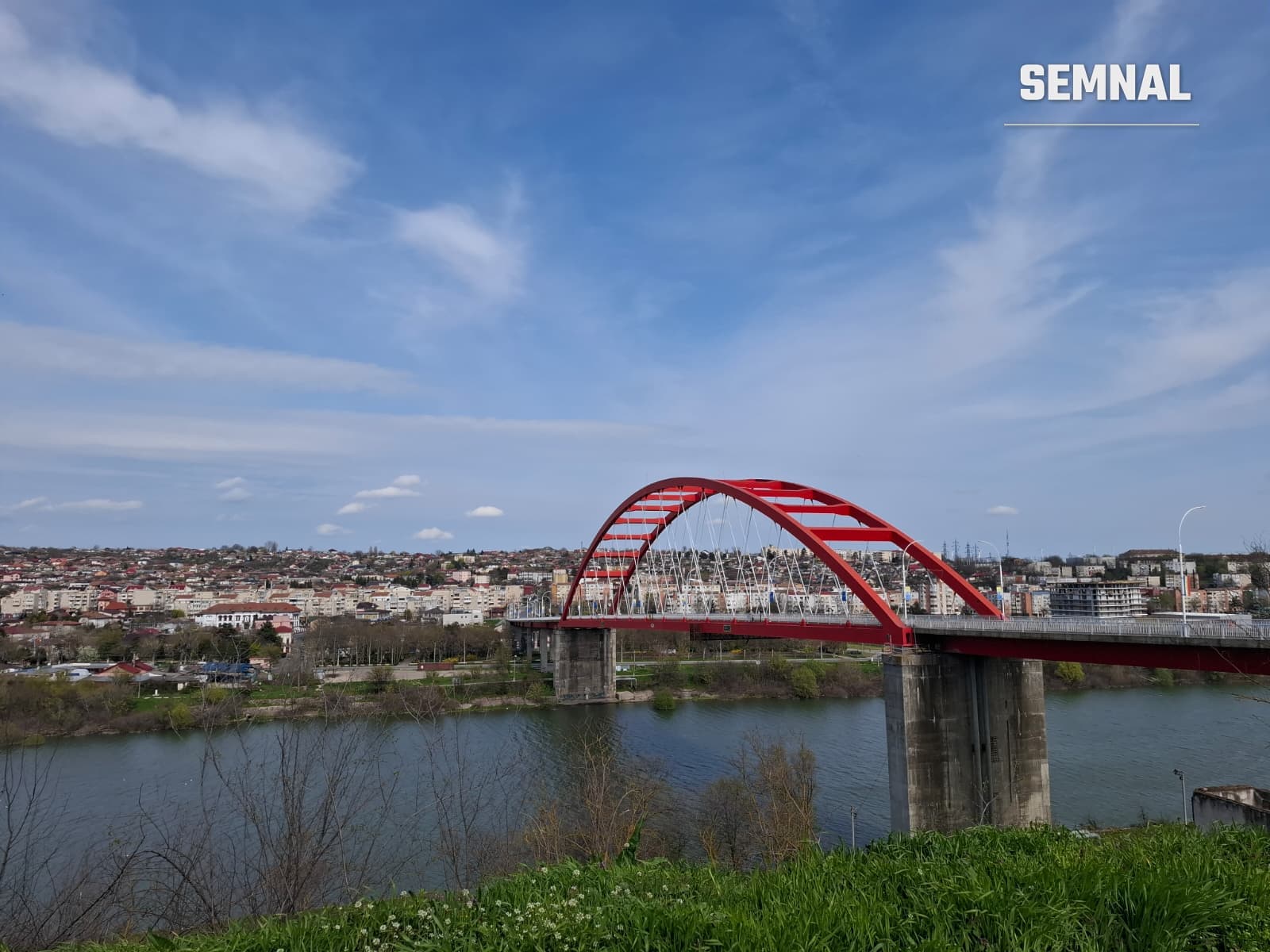 Pod metalic cu arc roșu peste un râu, cu clădiri de locuințe pe malul opus