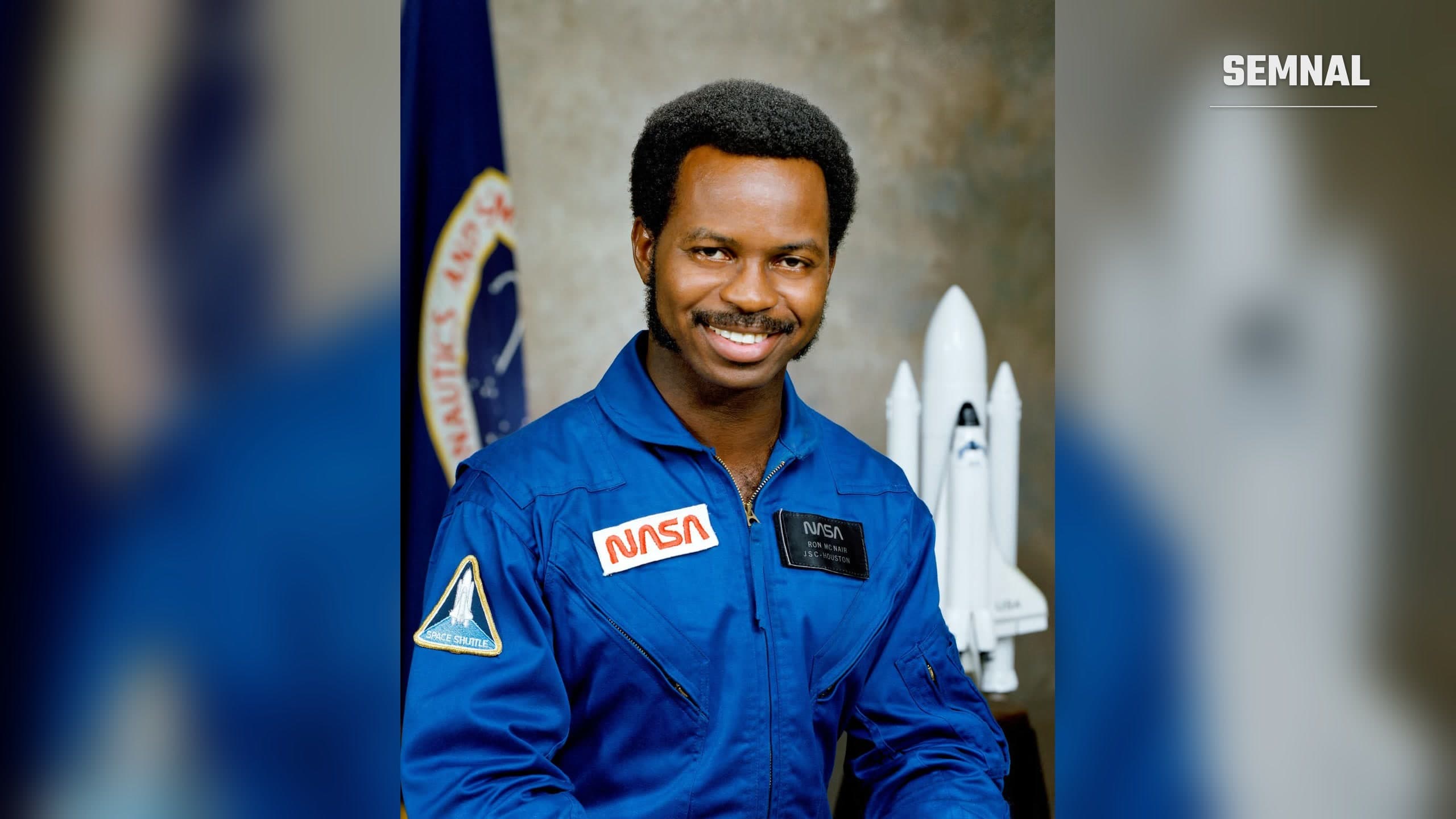 Portretul oficial NASA al astronautului Ronald McNair, în combinezon albastru, cu o machetă a navetei spațiale în fundal