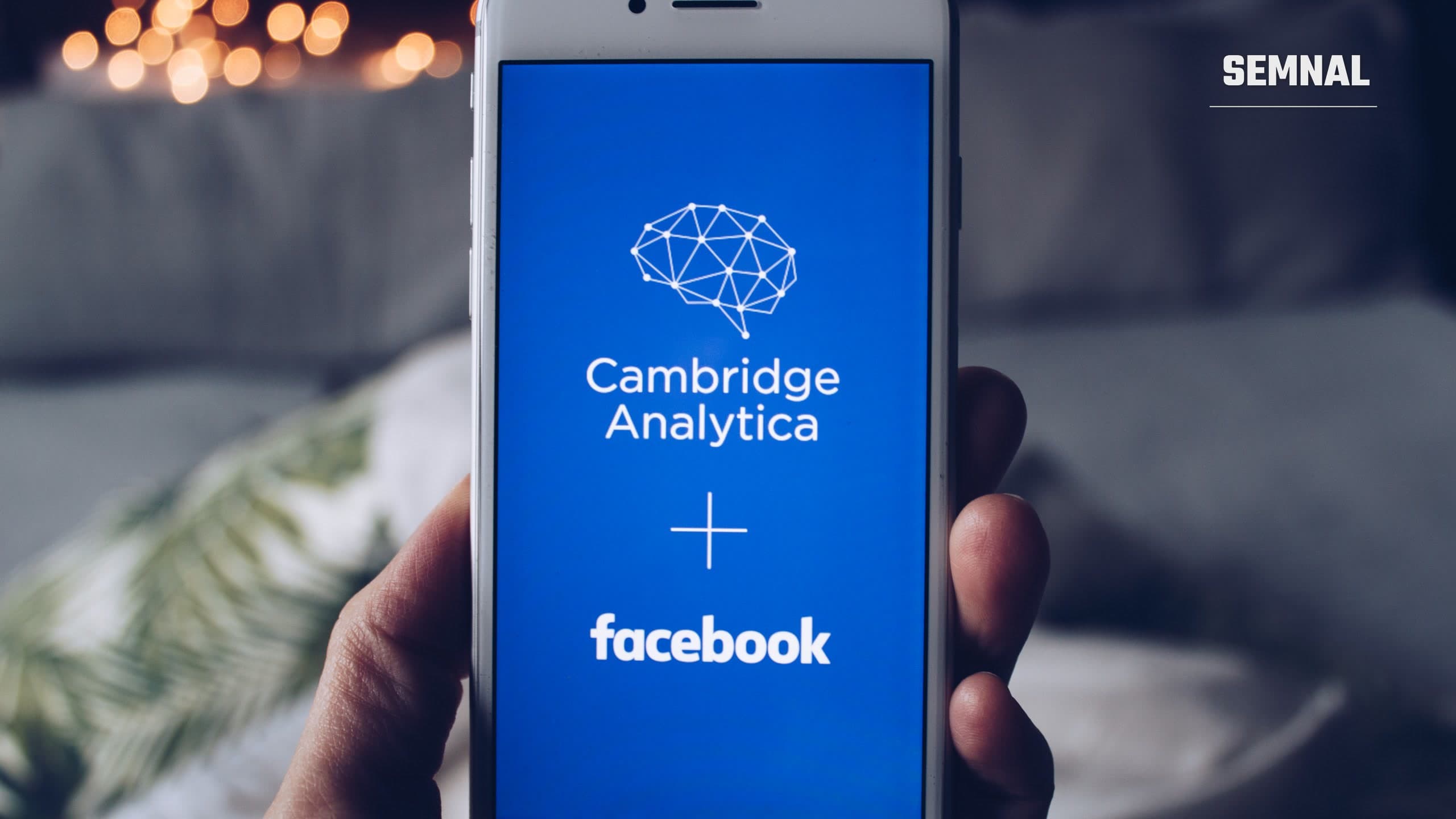 Telefon ținut în mână, cu ecranul albastru pe care apar siglele Cambridge Analytica și Facebook