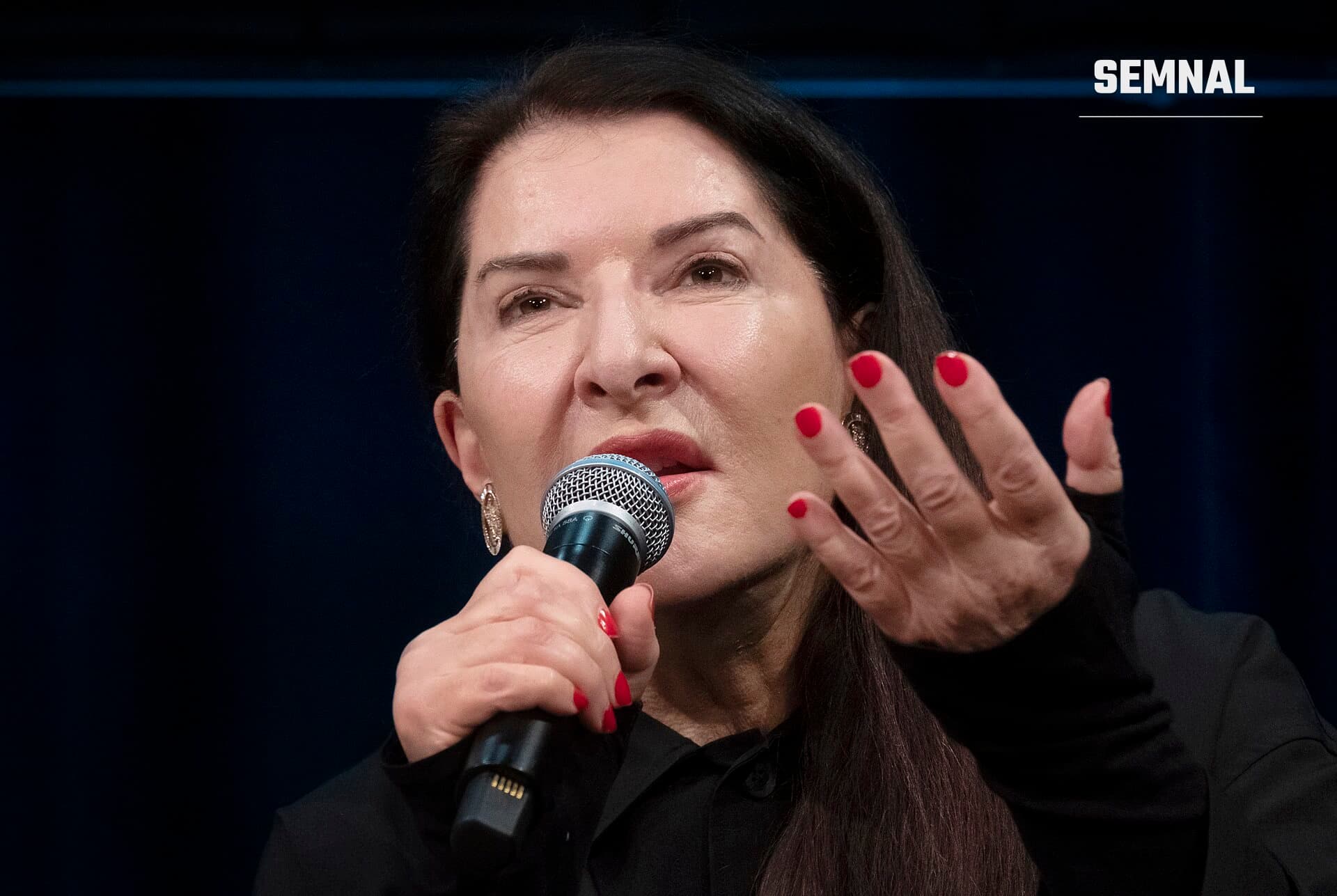Marina Abramović vorbind la microfon, cu mâna ridicată, la un eveniment din Amsterdam. (Foto: Anneheijs / Wikimedia Commons, CC BY-SA 4.0)