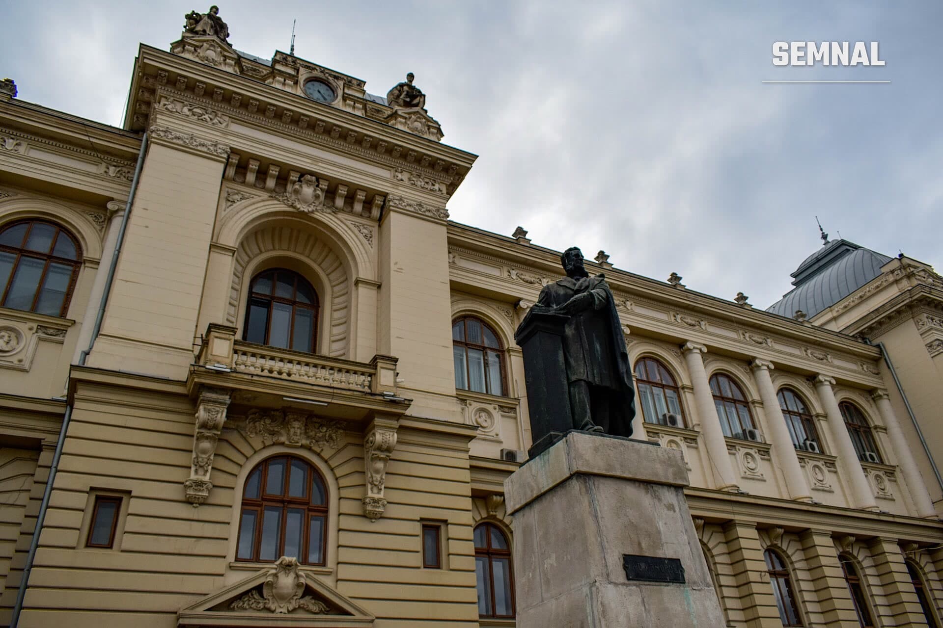 Clădirea Universității „Alexandru Ioan Cuza” din Iași, cu fațada crem și statuia din față, sub un cer înnorat