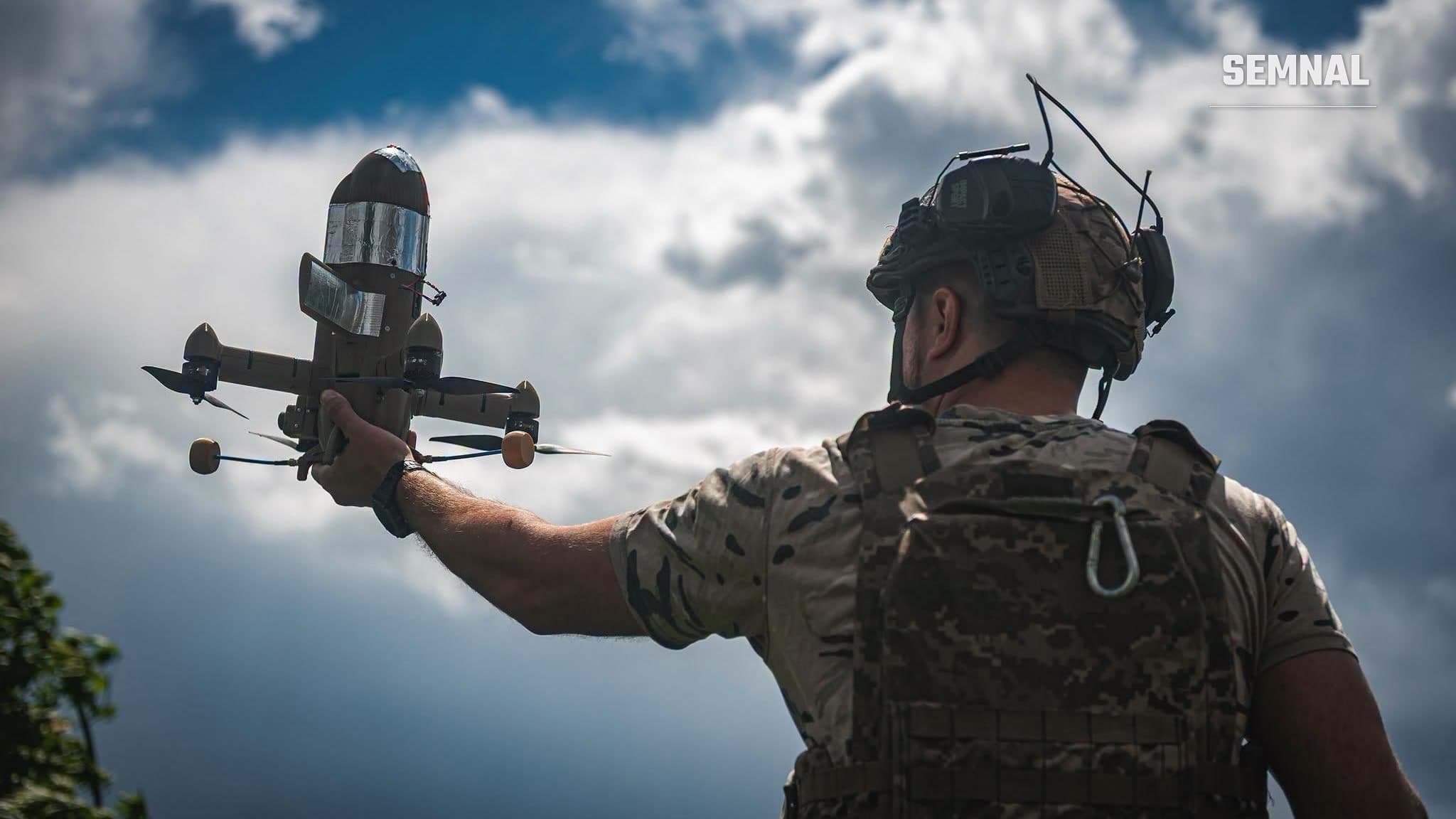 Un militar ucrainean în echipament de camuflaj, văzut din spate, ridică în mână o dronă mică, pe fundalul unui cer albastru cu nori