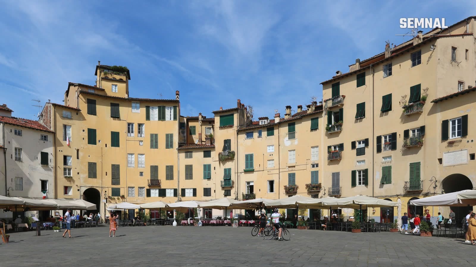 Clădiri galbene cu obloane verzi în jurul Piazza dell'Anfiteatro din Lucca, Toscana, cu turiști la mese și pe jos sub un cer albastru