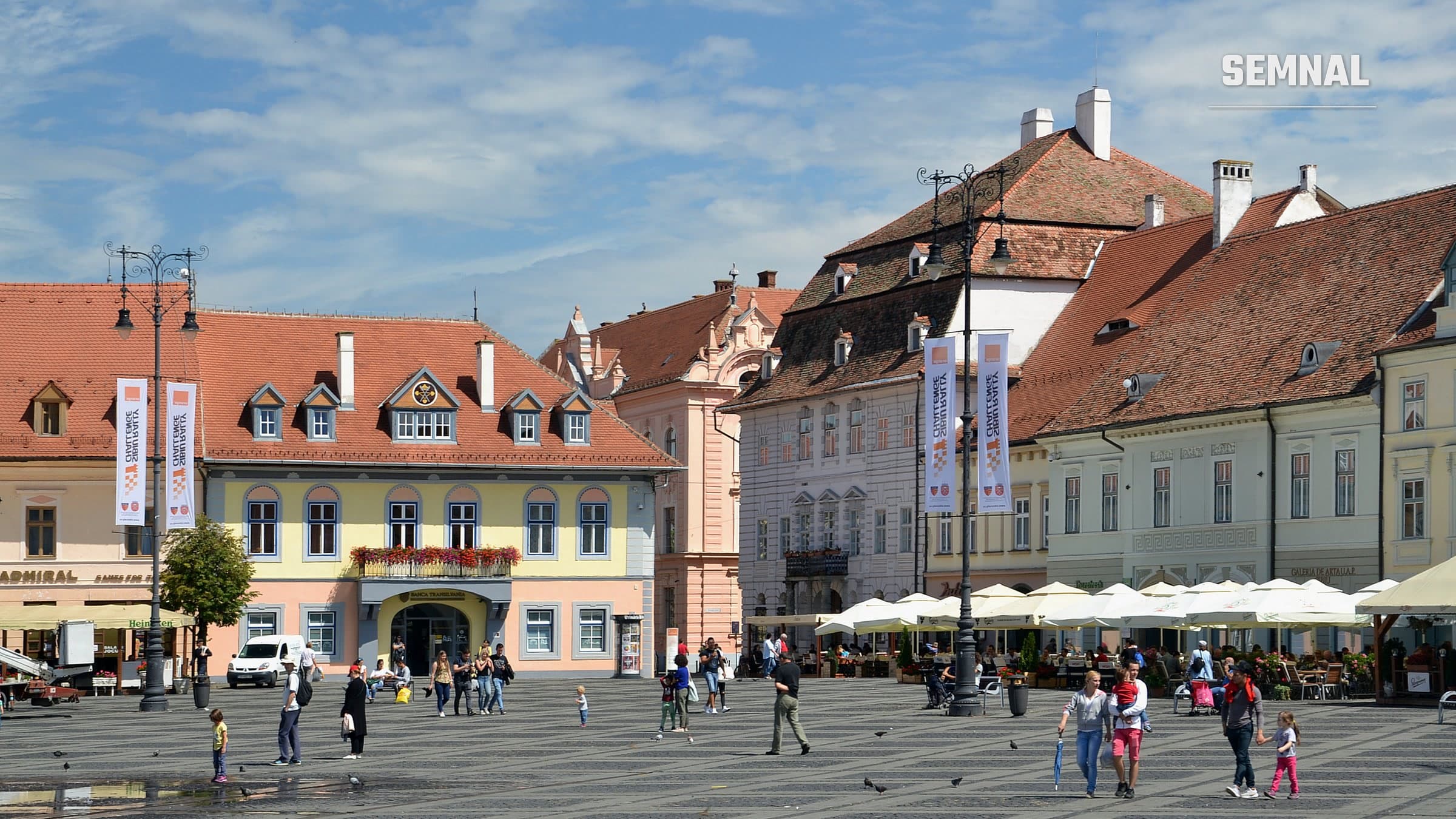 Turiști și localnici pe Piața Mare din Sibiu, într-o zi însorită, cu clădiri istorice cu acoperișuri de țiglă roșie și terase în fundal