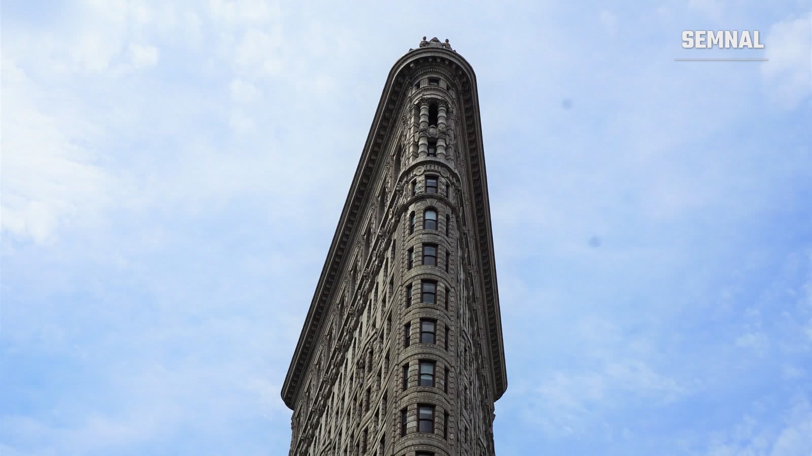 Vârful clădirii Flatiron din New York, cu fațada de piatră și cornișa, văzut de jos pe fundalul unui cer cu nori