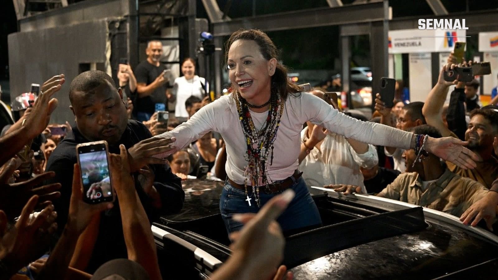 María Corina Machado cu brațele deschise, ieșită prin trapa unei mașini, înconjurată de susținători cu telefoanele ridicate, la Caracas