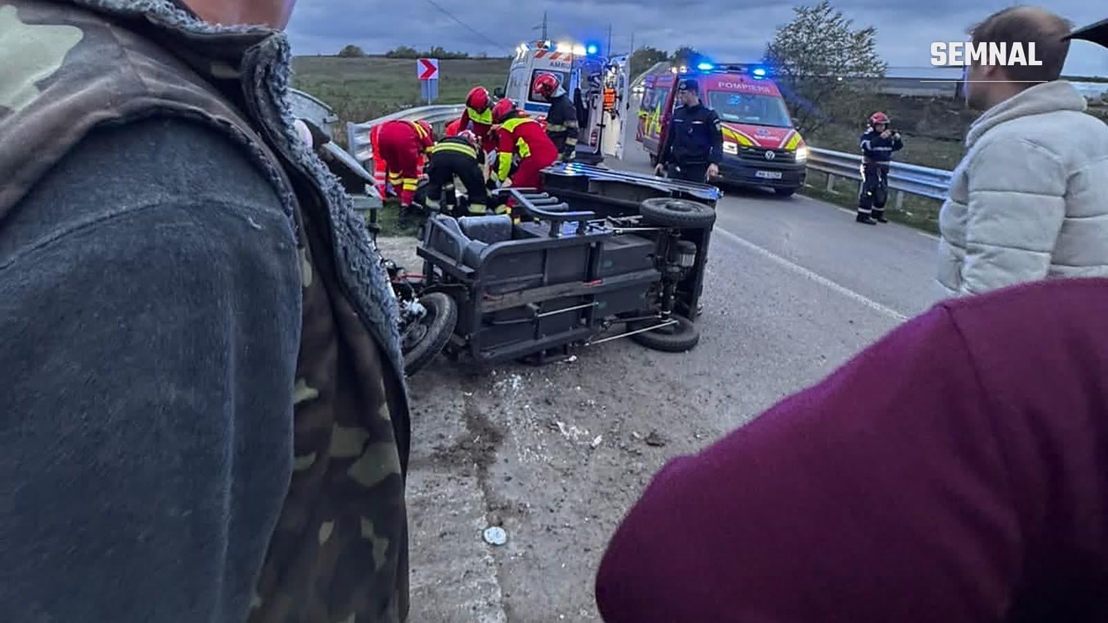 Vehicul cu trei roți răsturnat pe marginea unui drum, lângă un parapet, cu salvatori, o ambulanță și o autospecială de pompieri în fundal, seara