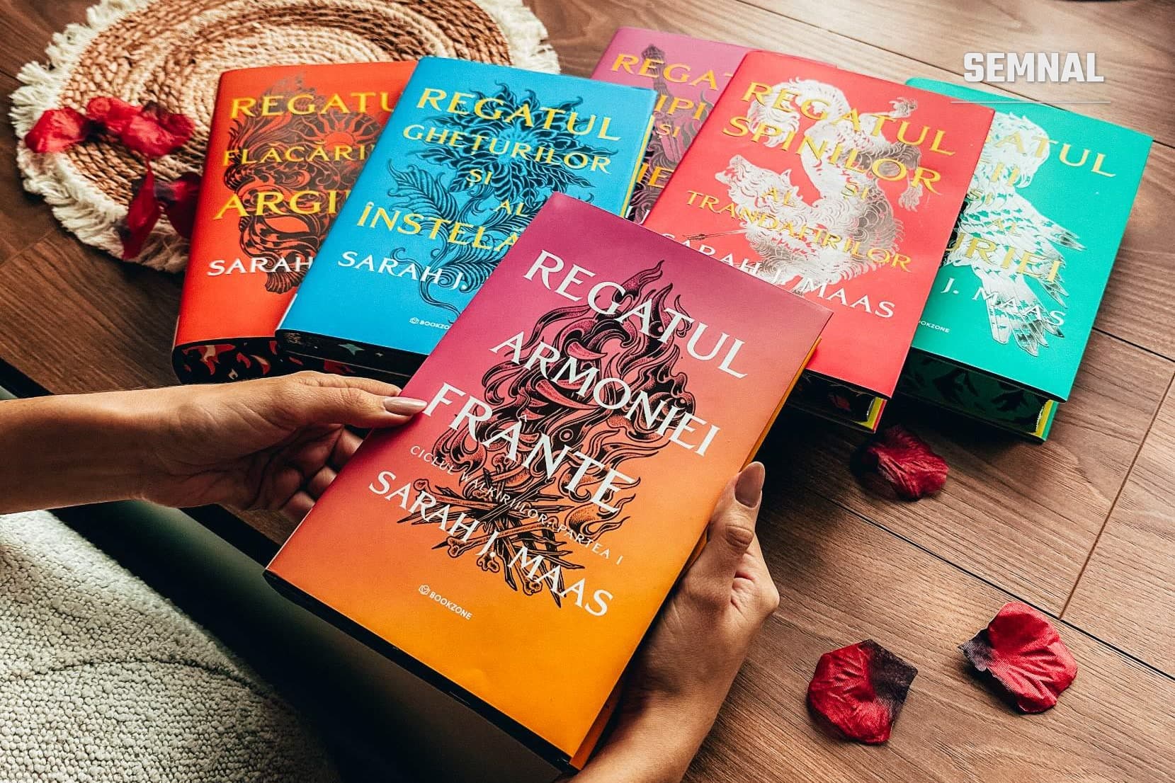 Mâini care țin volumul Regatul armoniei frânte de Sarah J. Maas, cu primele cinci volume ale seriei, în ediția Bookzone, așezate pe o masă de lemn