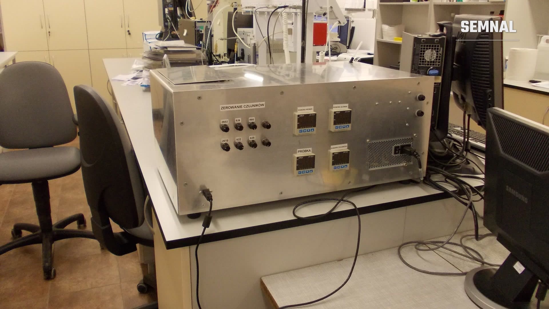 Prototip de nas electronic într-o carcasă metalică, pe un blat de laborator, cu butoane de reglare a senzorilor și afișaje digitale, lângă monitoare de computer