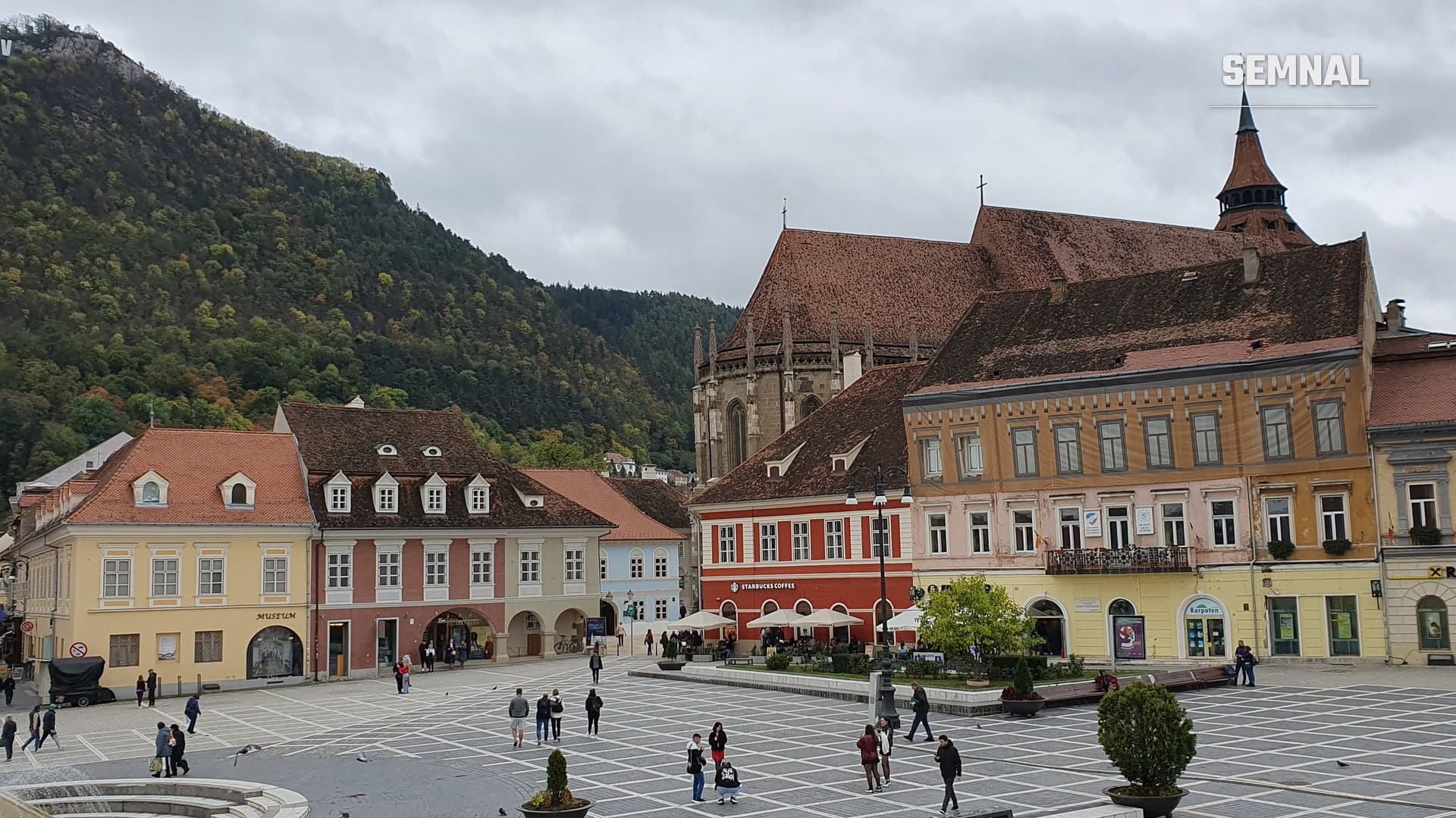 Turiști și localnici pe pavajul din Piața Sfatului din Brașov, cu Biserica Neagră și dealul Tâmpa în fundal, într-o zi de toamnă înnorată