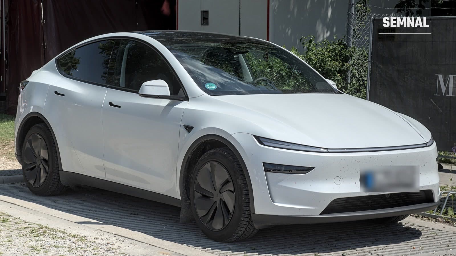 Tesla Model Y alb, văzut din față și din lateral, parcat pe un pavaj cu dale perforate, lângă un gard, cu numărul de înmatriculare estompat