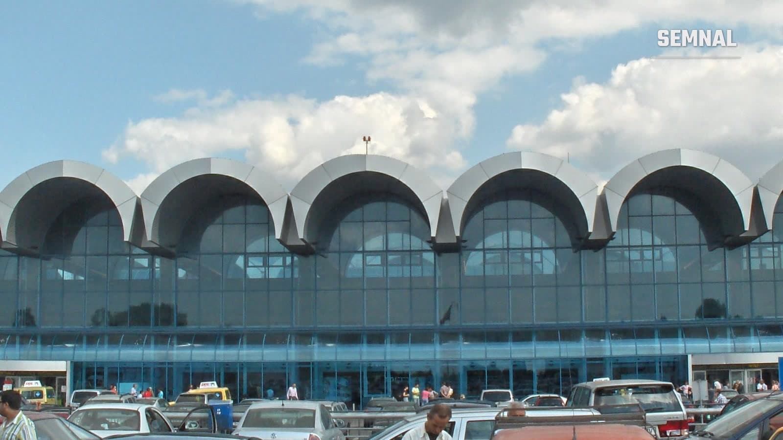 Clădirea Terminalului Sosiri a Aeroportului Internațional Henri Coandă din Otopeni, cu fațada din sticlă și arcade, văzută din parcare
