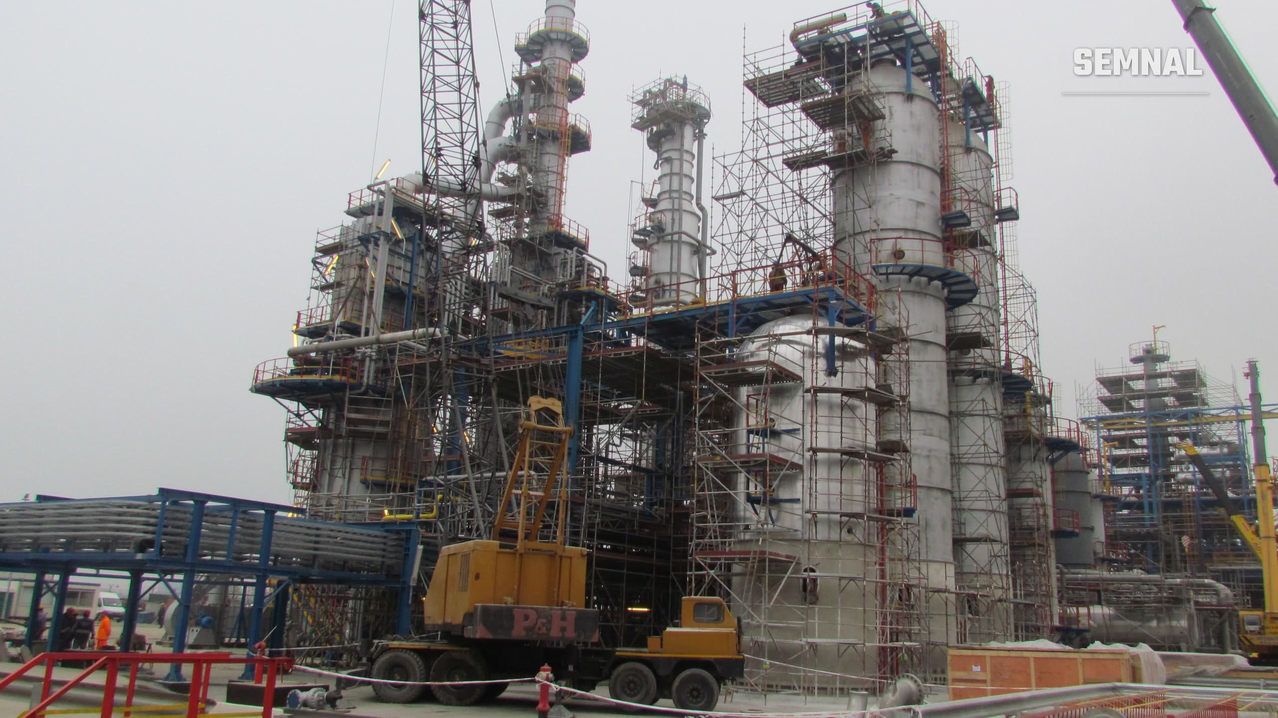 Instalații ale rafinăriei Petromidia din Năvodari: coloane metalice de proces înconjurate de schele, cu o macara galbenă și muncitori în prim-plan, sub un cer cenușiu