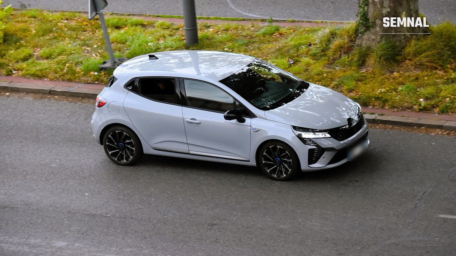 Renault Clio E-Tech hybrid gri deschis, văzut din lateral-față și de sus, în mers pe o stradă din Franța, lângă o bordură cu iarbă