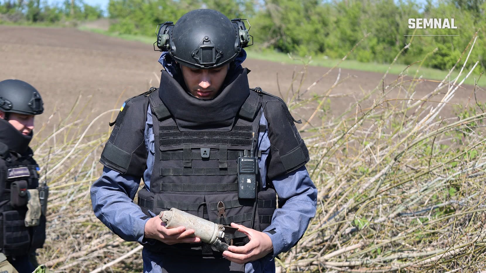Doi deminatori ai Serviciului de Urgență al Ucrainei, în căști și veste de protecție, într-un câmp; unul examinează o muniție. Fotografie ilustrativă din regiunea Harkov.