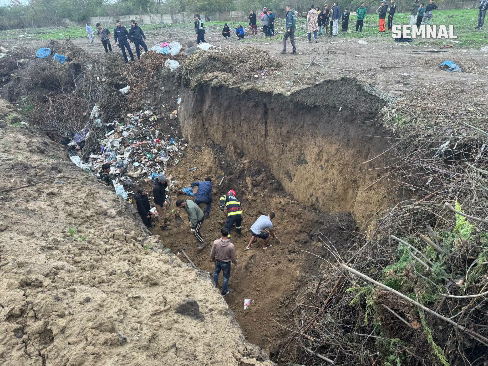 Un pompier și mai multe persoane în haine civile sapă cu lopeți în fundul unei gropi adânci, iar polițiști și localnici privesc de pe marginea ei