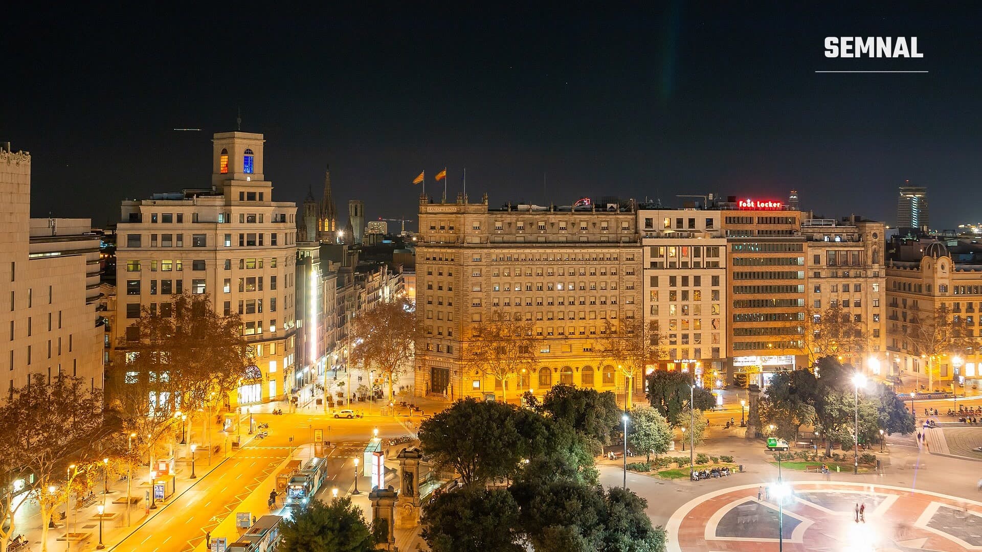 Vedere nocturnă asupra pieței Plaça de Catalunya din Barcelona, cu clădiri luminate și o intersecție cu autobuze. Foto: MB-one / Wikimedia Commons, CC BY-SA 4.0, decupată.