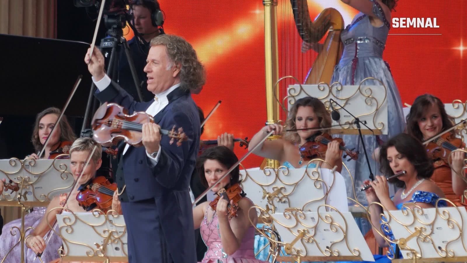 André Rieu cântă la vioară pe scenă, în fața muzicienelor Orchestrei Johann Strauss îmbrăcate în rochii de bal