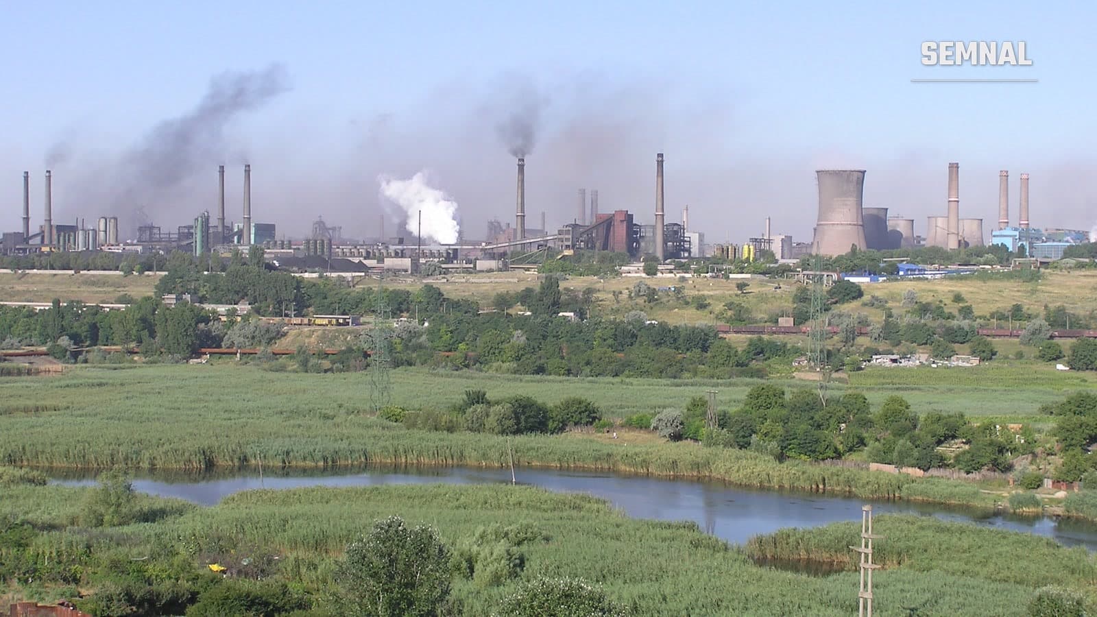 Panoramă a combinatului siderurgic de la Galați, cu coșuri de fum și turnuri de răcire, văzută peste o zonă cu stuf și un canal