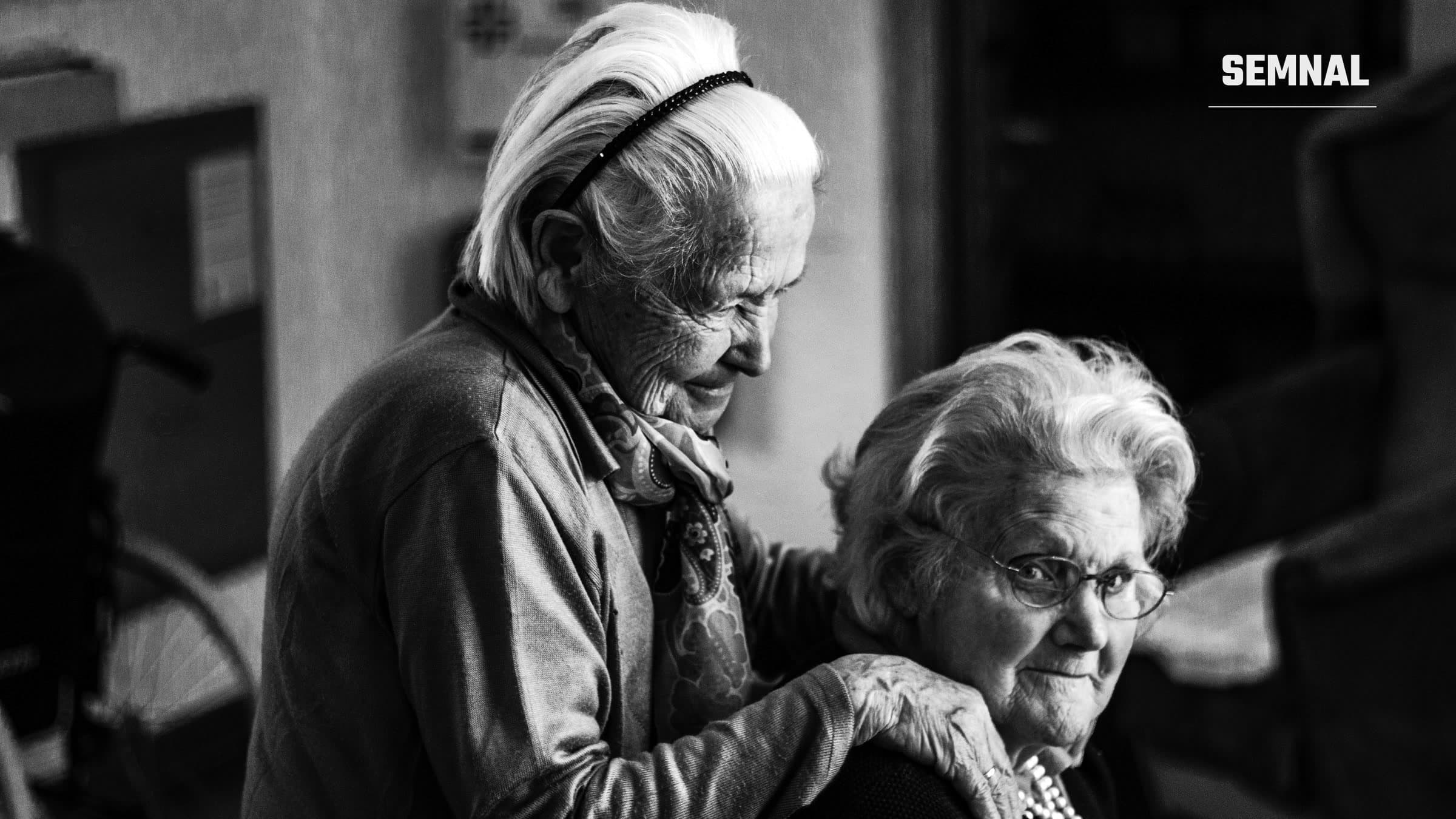 Două femei în vârstă, cu părul alb, într-o cameră: una stă în spate și își sprijină mâinile pe umerii celeilalte, care privește spre cameră