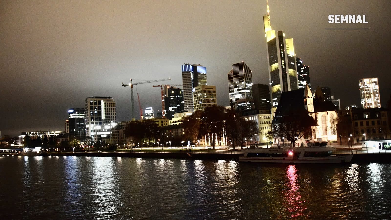 Silueta zgârie-norilor din Frankfurt luminați noaptea, văzută peste râul Main, cu o biserică veche și un vas de agrement pe mal