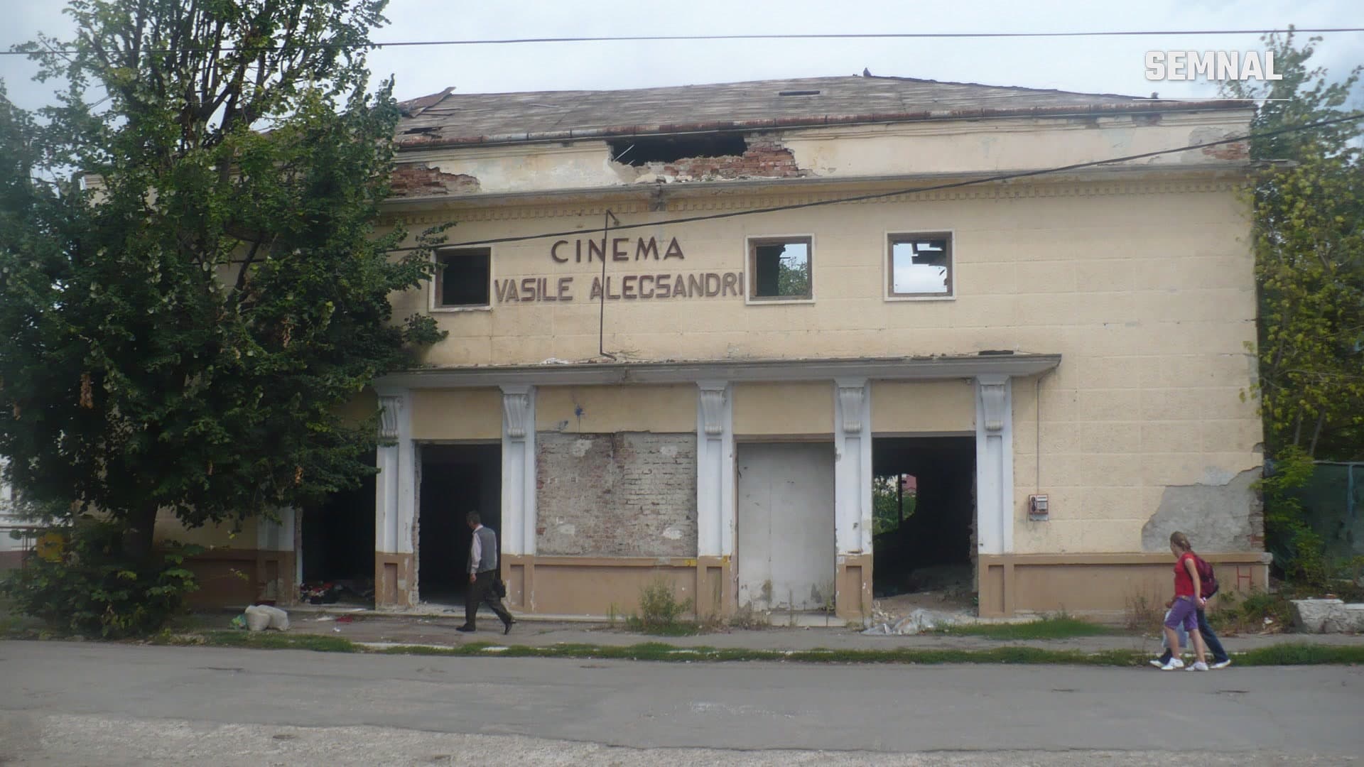 Fațada degradată a cinematografului Vasile Alecsandri din Roman, cu geamuri sparte, acoperiș deteriorat și intrări închise, în timp ce doi trecători merg pe trotuar