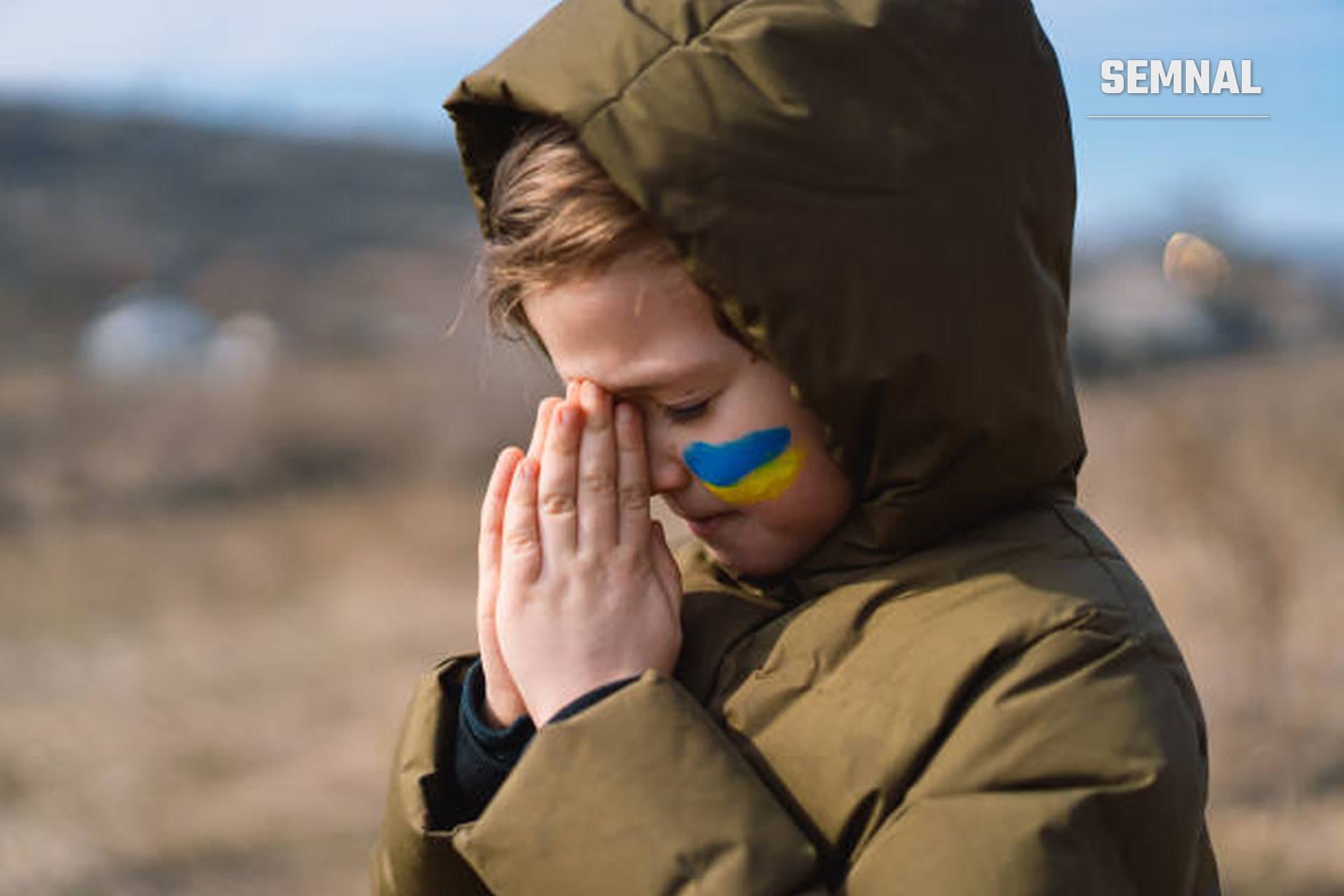 În imagine este un copil din Ucraina care se roagă pentru pace. Are pe unul din obraji pictate culorile steagului țării lui: galben și albastru.