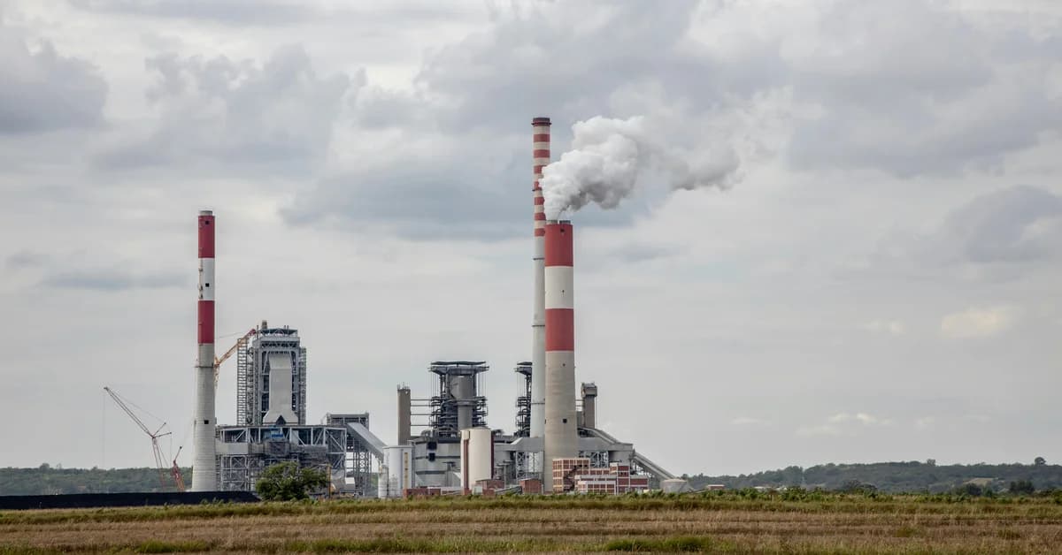O fabrică industrială cu coșuri de fum mari eliberând abur sau fum, înconjurată de vegetație și cer înnorat.