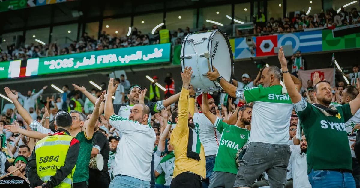 Fani sirieni sărbătorind în tribună, îmbrăcați în tricouri albe și verzi, cu steaguri și tobe, într-un stadion aglomerat.