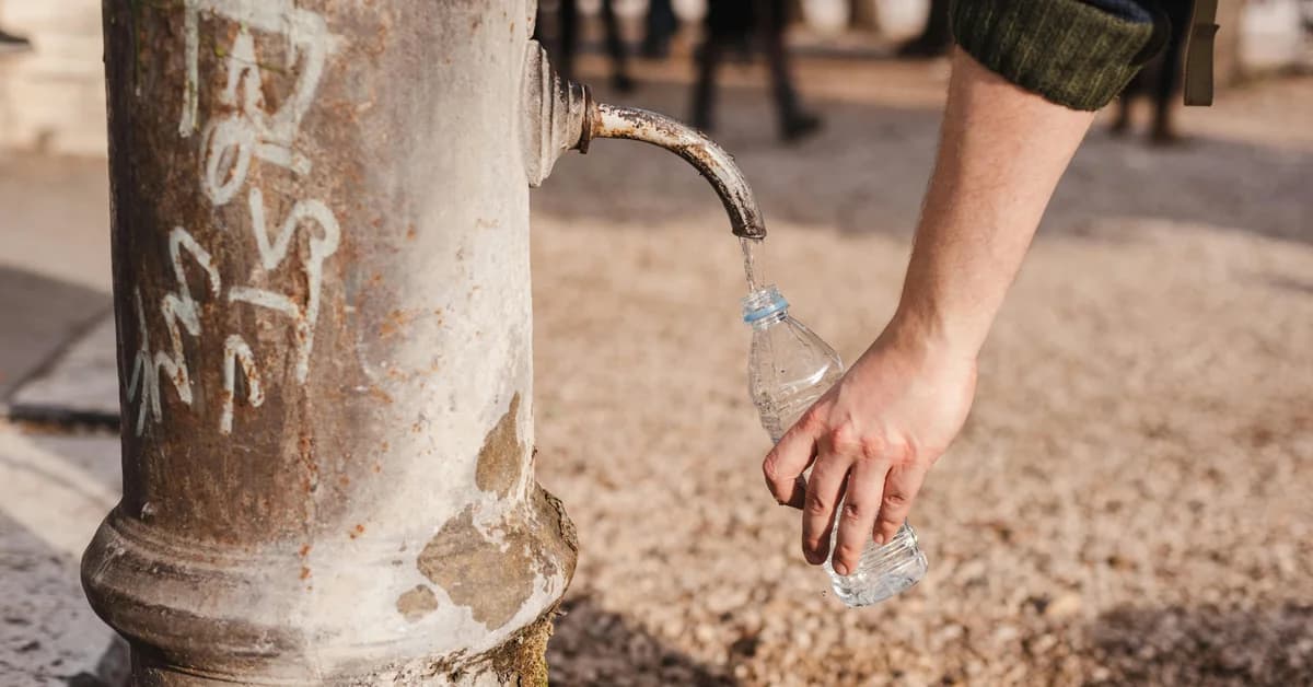 Un robinet de apă vechi cu graffiti și o persoană care umple o sticlă de plastic cu apă, în aer liber.