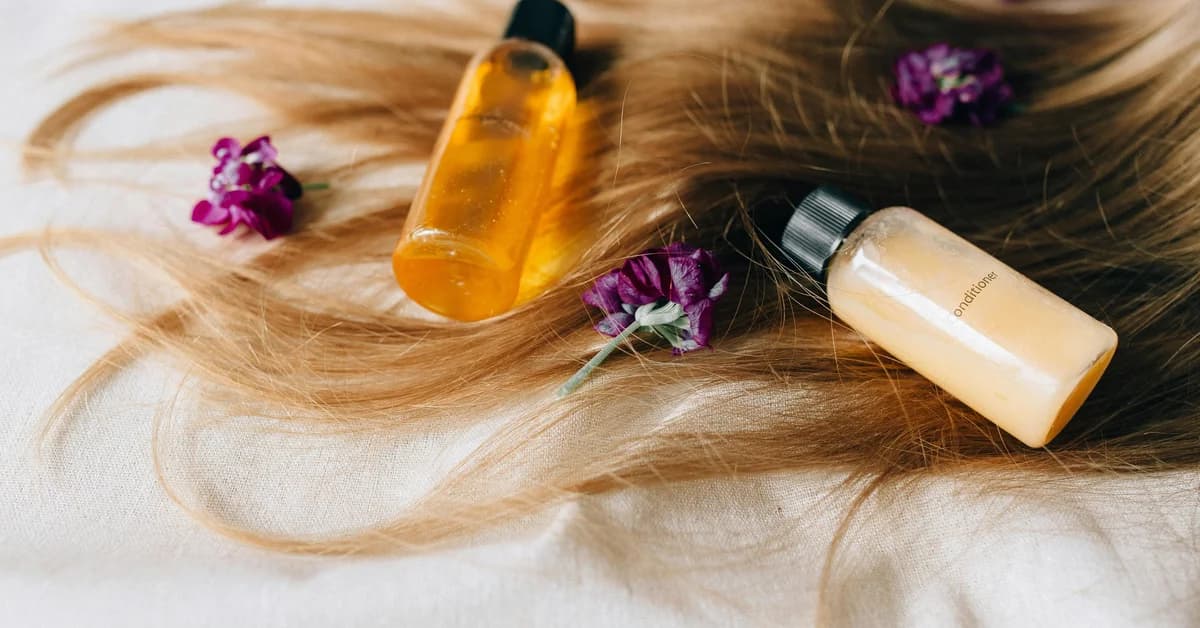 Flacoane cu esență pentru păr pe păr blond desfăcut, alături de flori mov pe o suprafață albă.