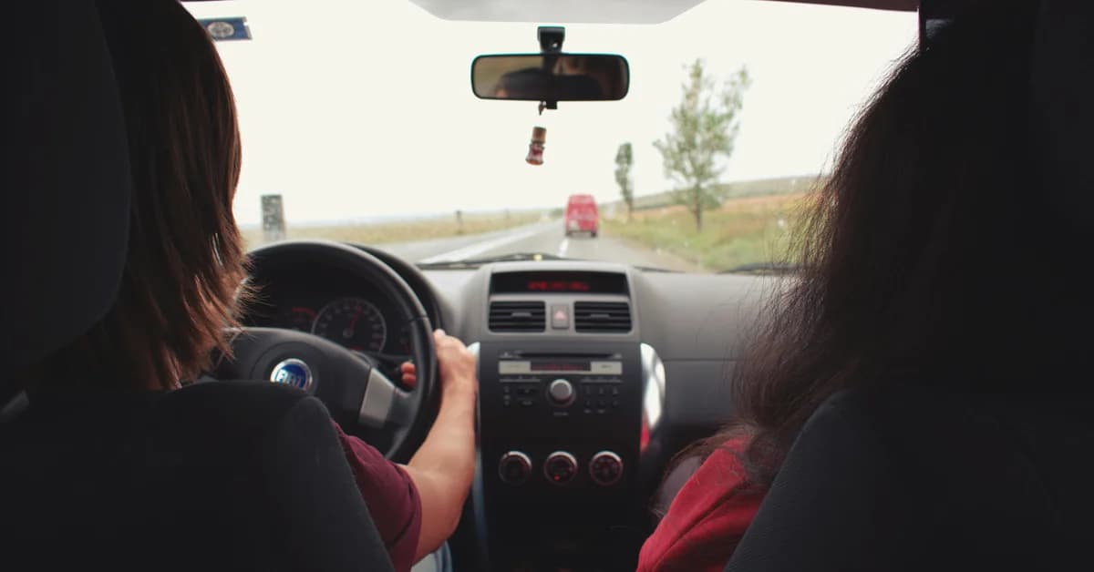 Un instructor auto și un elev într-o mașină, pe un drum rural, cu un copac și un vehicul roșu în față.