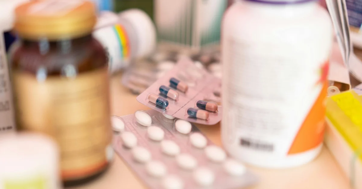 Imagine cu blistere și flacoane de medicamente pe o masă, unele fiind neclare în fundal.