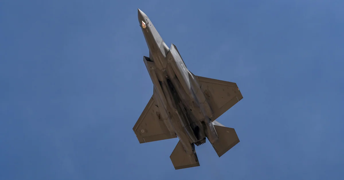 Avion de luptă zburând pe cer, surprins de jos, cu o parte din nori în fundal.