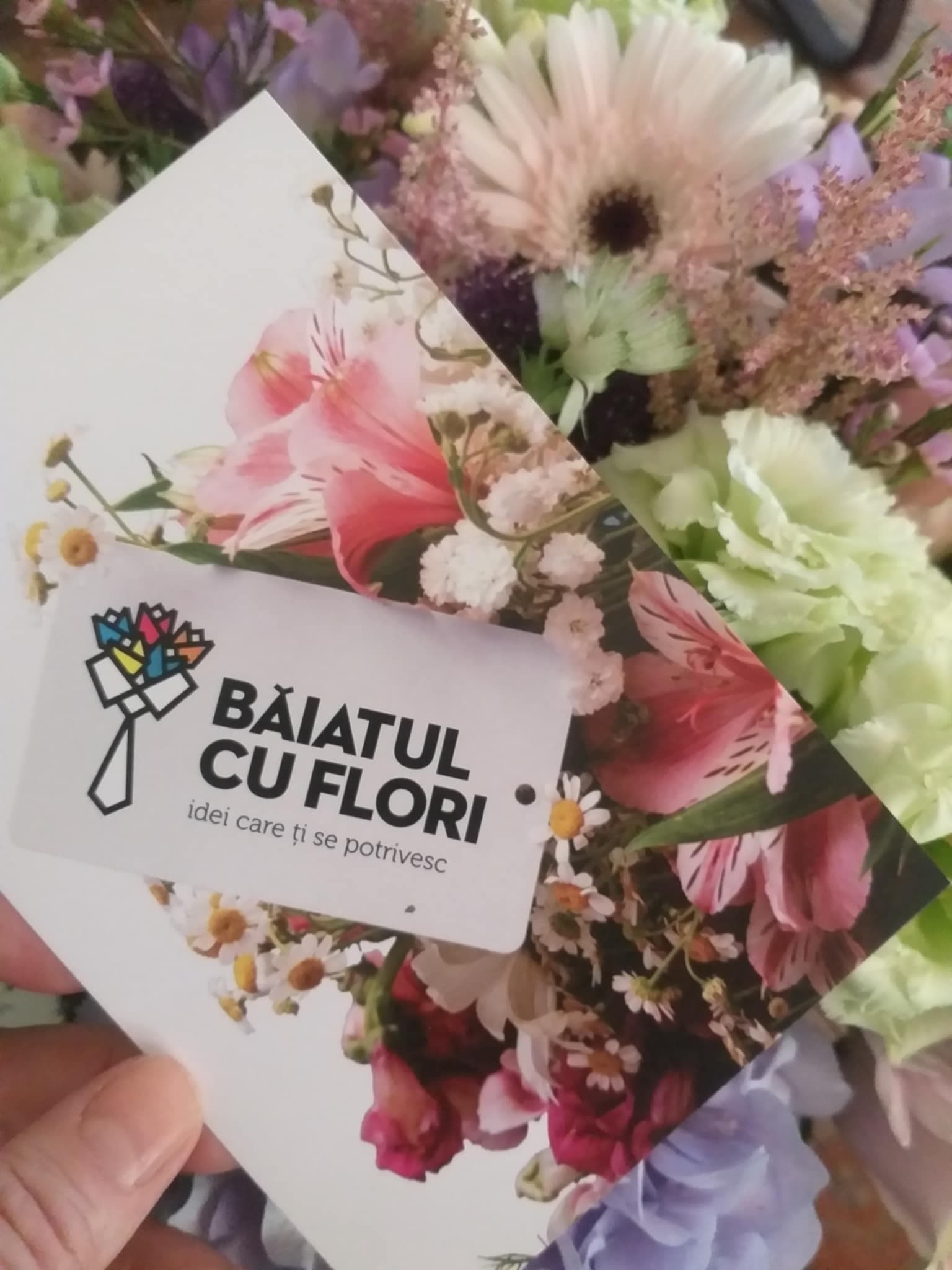 Mână ținând etichetă albă cu text 'Baiatul cu flori - idei care ți se potrives' deasupra unui buchet cu trandafiri roz, gerbere albe și flori mov