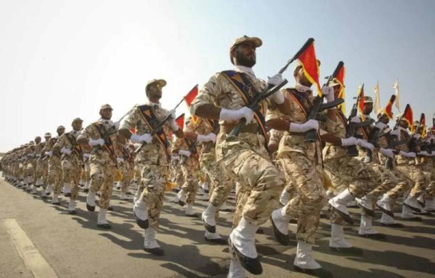 Soldații în uniforme de desert marșează în parada militară, purtând steaguri iraniene și arme, formând o linie compactă pe o cale asfaltată sub soare intens.