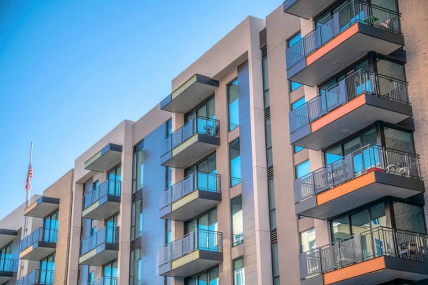 Bloc modern de locuințe cu mai multe etaje, fațadă în tonuri deschise, ferestre mari și balcoane cu balustrade metalice negre și accente portocalii sub cer senin.