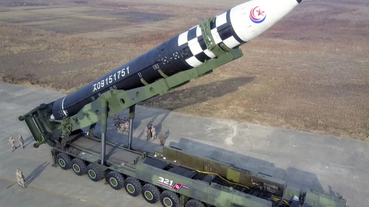 Sistem de lansare de rachete balistice nord-coreene de tip Hwasong pe platformă militară, cu coiful alb și emblemă națională, în teren muntos.