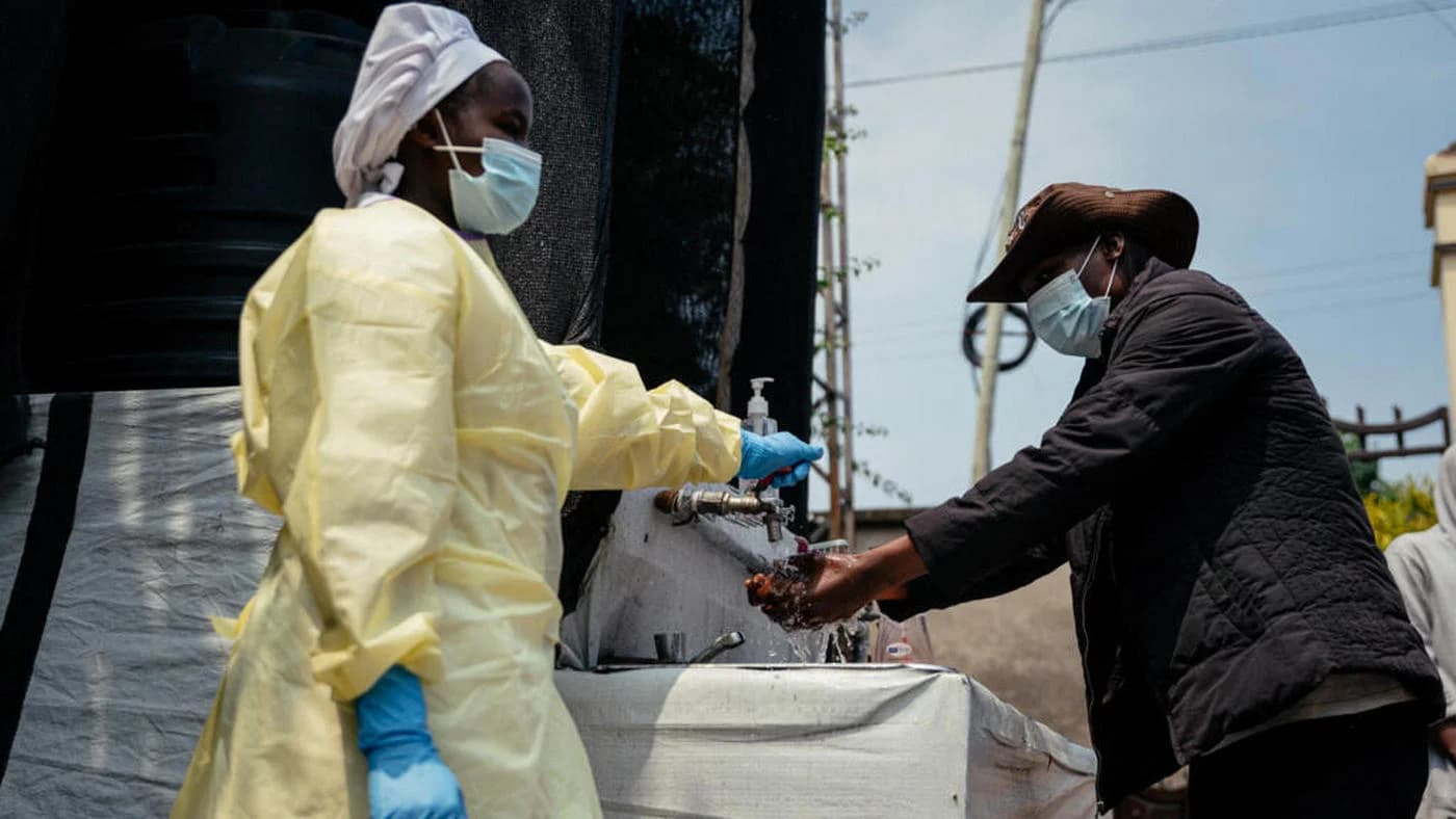 Doi lucrători în echipament de protecție cu costume galbene și măști medicale dezinfectează un vas cu apă, part al eforturilor de control epidemiologic în Congo pentru combaterea focarelor de Ebola.