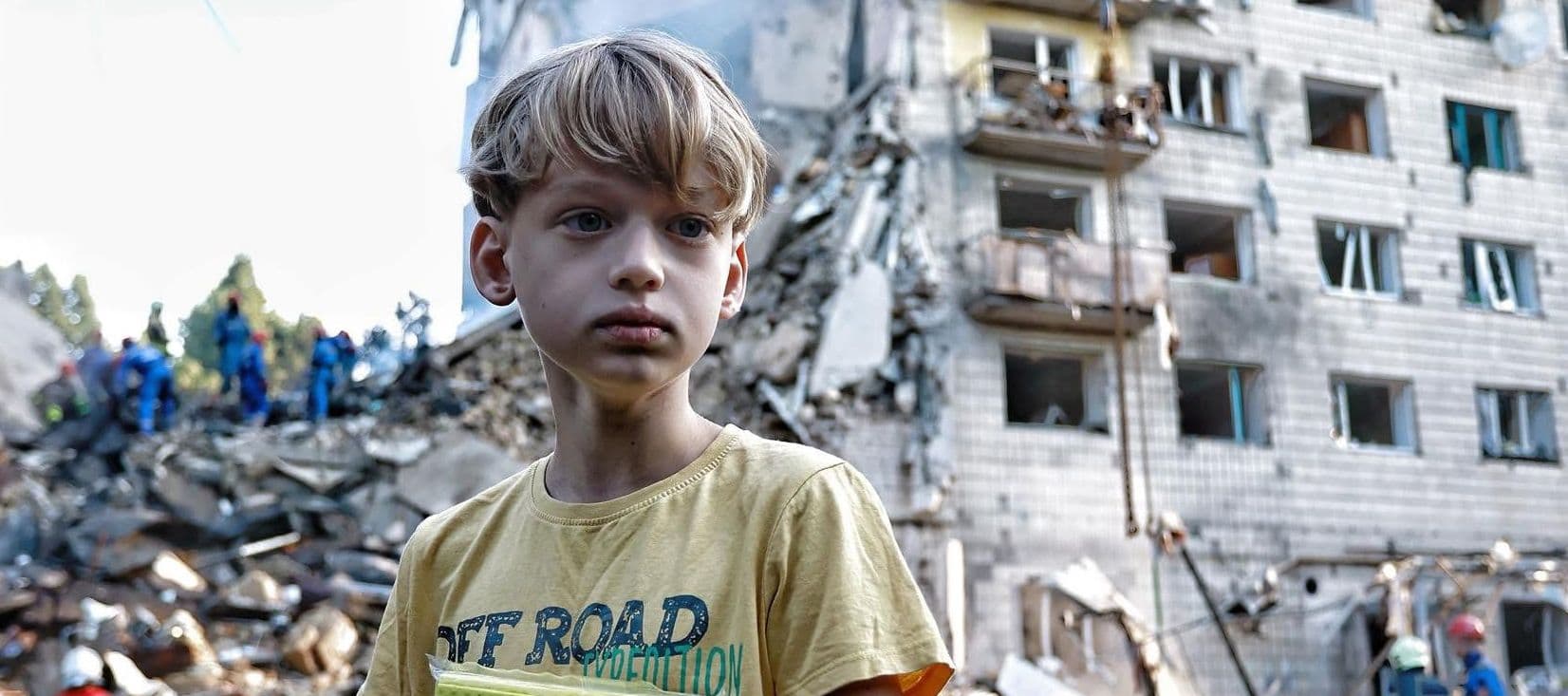 Copil cu părul blond în fața unei clădiri cu daunele din atac aerian, moloz și echipe de salvare în fundal la Kiev.