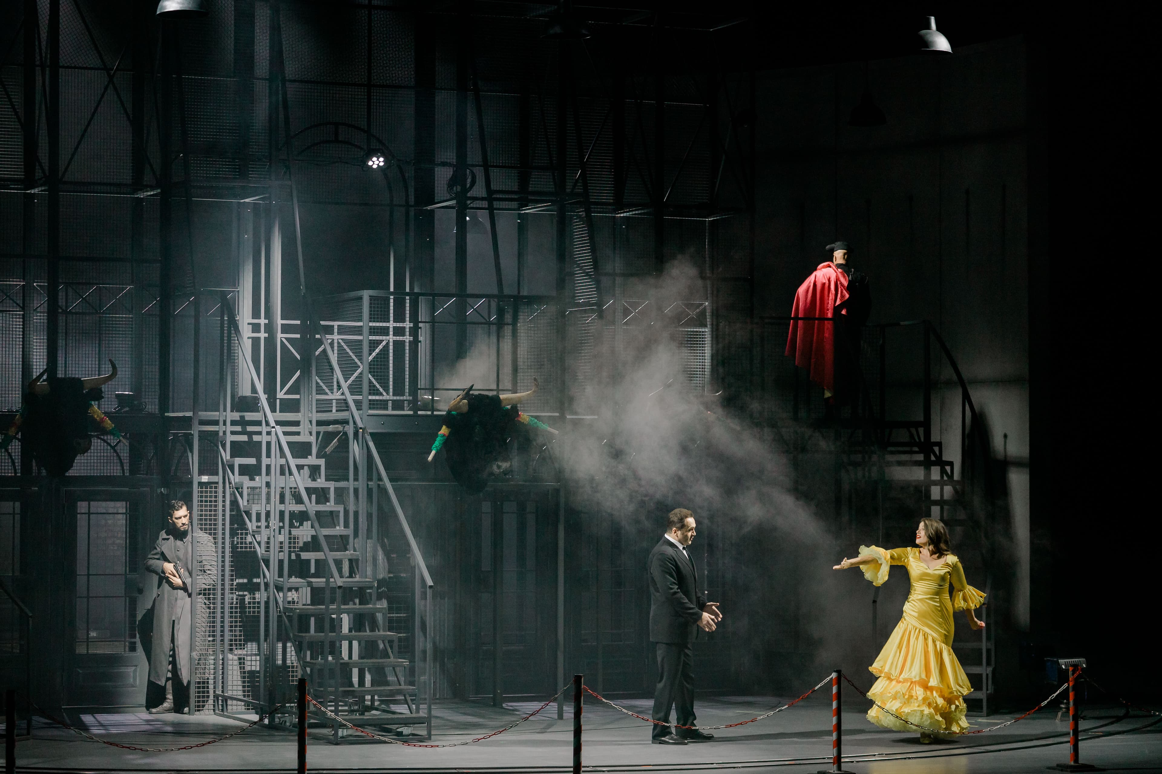 Scenă din spectacolul „Carmen” la Opera Națională București, cu un bărbat în costum și o femeie în rochie galbenă pe o scenografie metalică