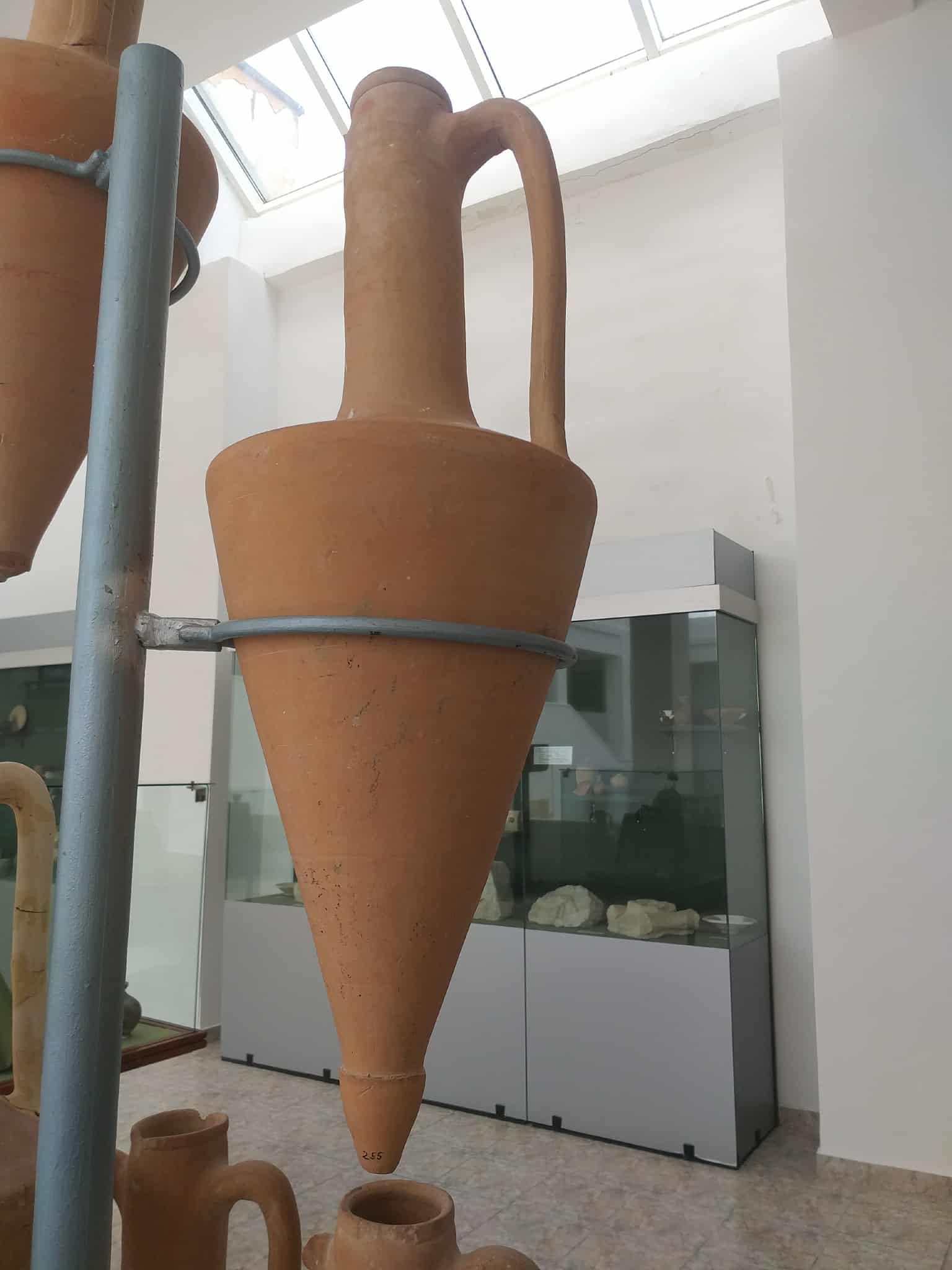 Amforă antică de teracotă cu formă conică și două mânere, exponată în muzeu pe suport metalic, cu vitrine în fundal