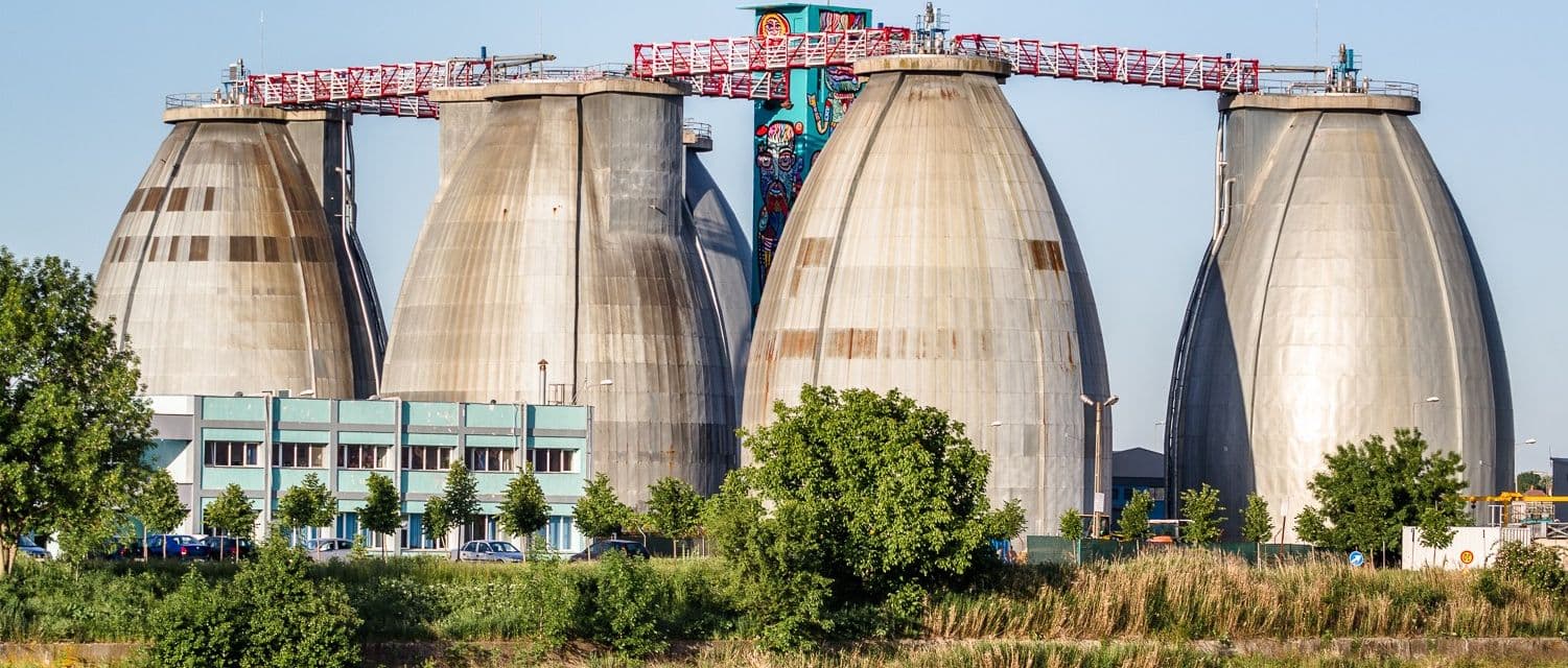 Stație de epurare a apei uzate cu patru rezervoare conice din beton, macara de ridicare și clădire de control, pe fundalul unui pajiș verde.