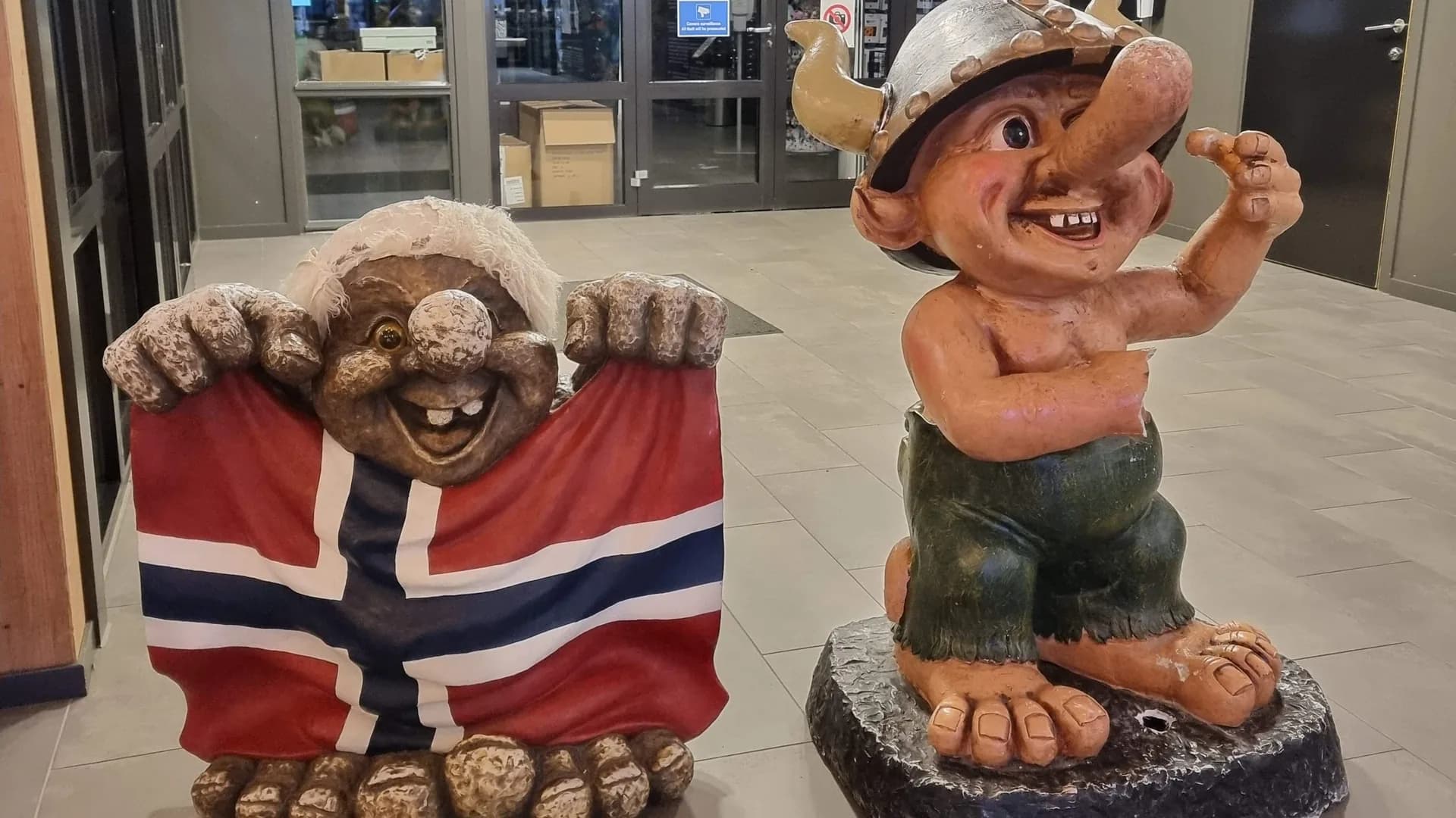 Două statuete de troli norvegieni la intrarea unui mall, unul ținând drapelul Norvegiei, celălalt cu un coif cu coarne.