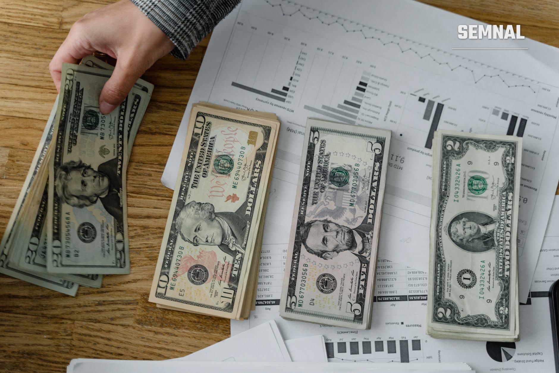 Mână ținând bancnote de dolar american pe masa cu documente financiare și grafice; ilustrând tranzacții și sume mari în context de negocieri economice.