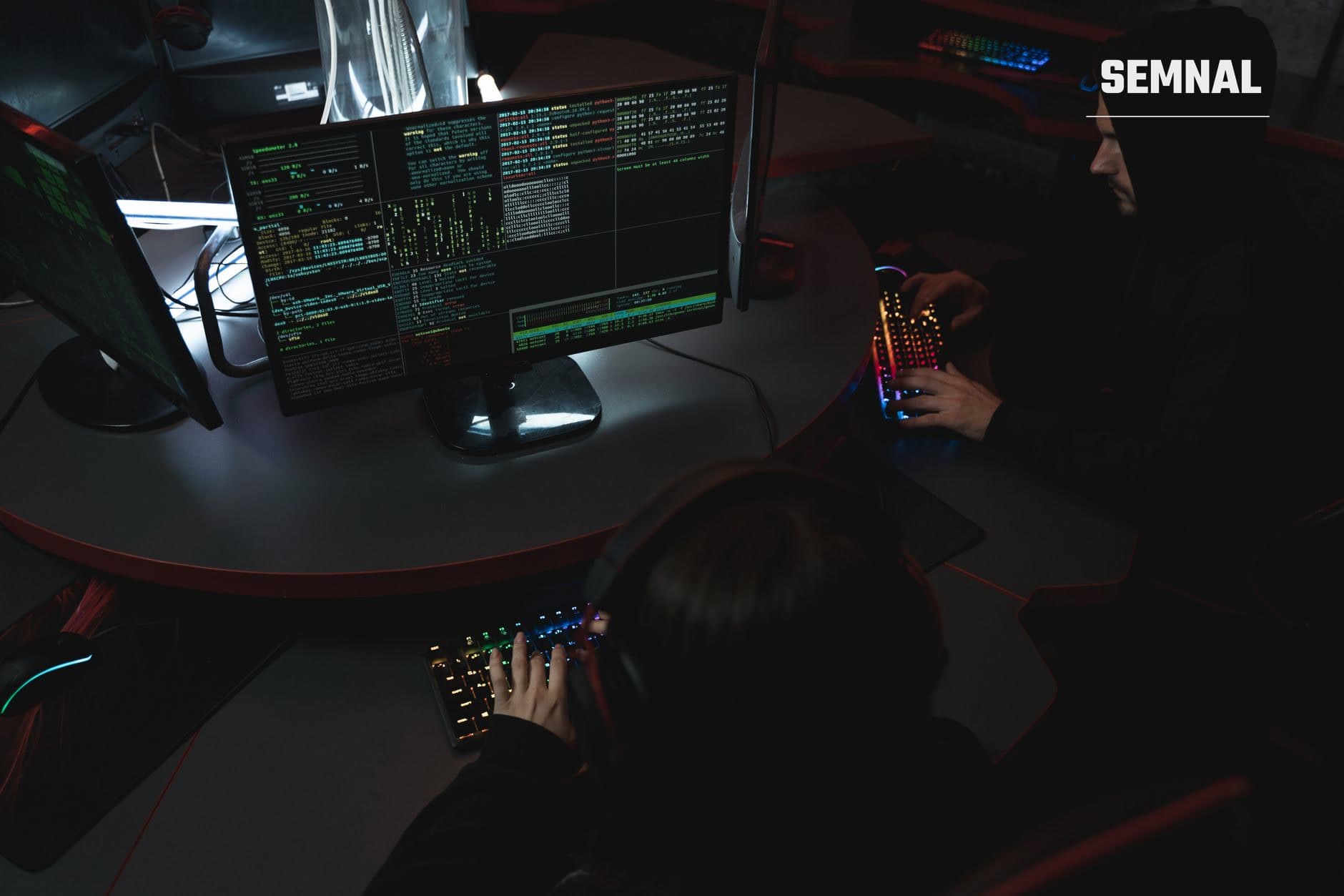 Doi hackeri lucrând la calculatoare cu cod pe ecrane și tastaturi mecanice iluminate în laborator de securitate cibernetică.