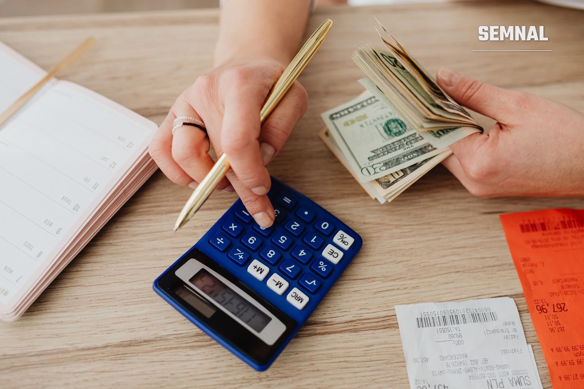 Persoană calculând cheltuieli lunare cu un calculator albastru, ținând bancnote și documente financiare pe masă pentru organizarea bugetului familial