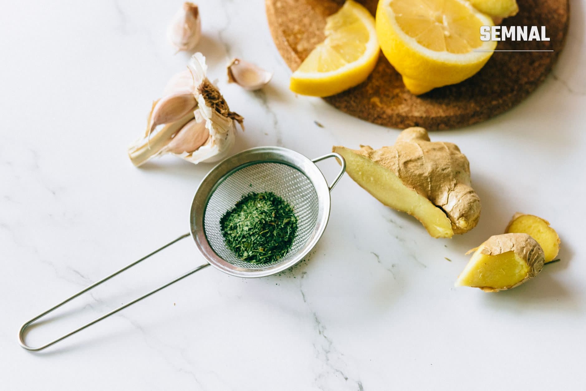 Ingrediente naturale pentru imunitate: lămâie tăiată, usturoi, ghimbir proaspăt și ceai verde în sită, pe blat alb de marmură