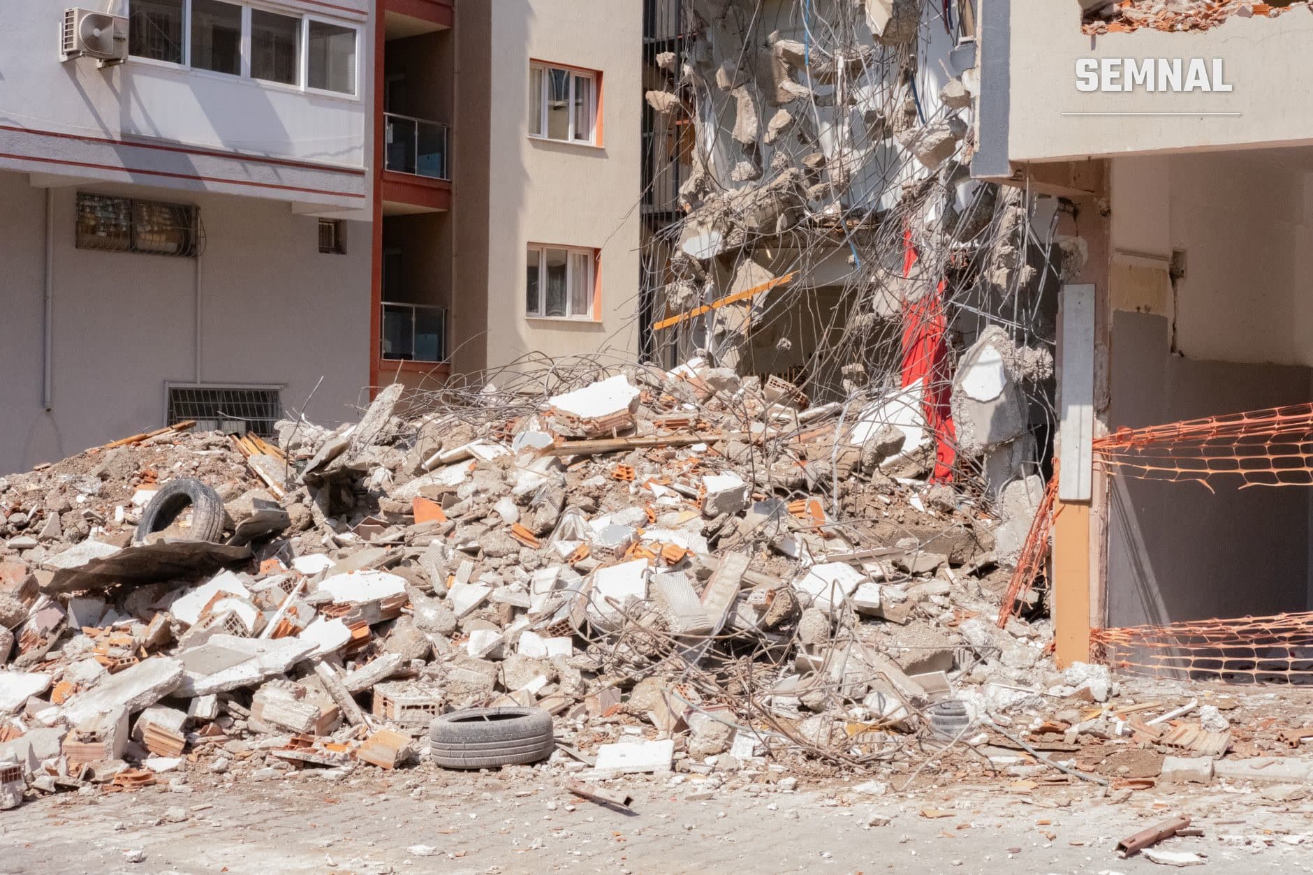 Ruine ale unui bloc de locuințe distrus de cutremur, cu moloz de ciment și cărămidă răspândit, grinzi metalice roșii expuse și fire electrice întortocheate într-o curte între clădiri dăunate.