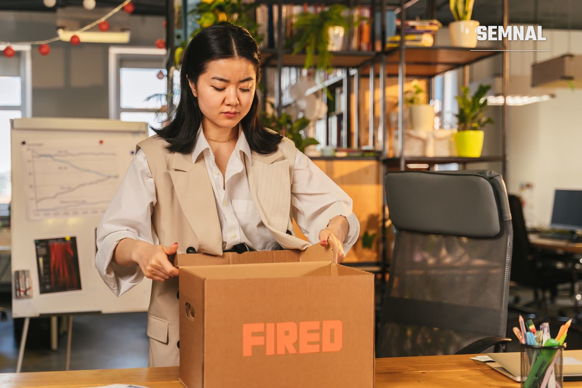 O angajată ține o cutie de carton marcată "FIRED" într-un birou modern, reprezentând încheierea unei relații de muncă și procesul de demisie sau concediere.
